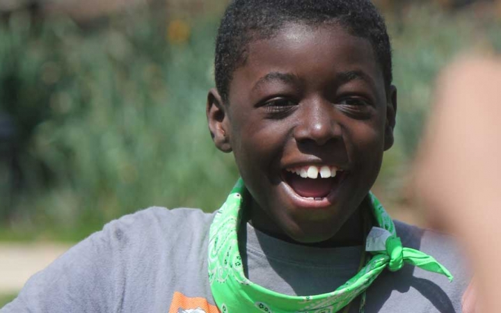 A young person smiles a big smile. There is blurry greenery in the background. 